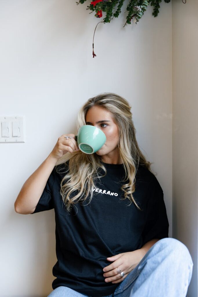 A young woman drinking from a mug, seated in a relaxed indoor environment.